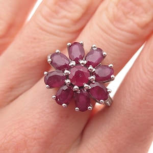 May include: A silver-toned ring featuring a central round ruby surrounded by eight oval-cut rubies arranged in a floral pattern. The deep red gemstones are set in metal prongs, creating a cluster design that resembles a flower blossom on the finger.