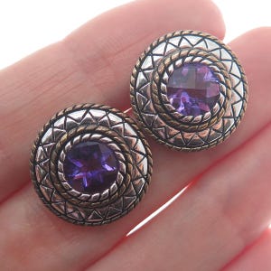 May include: A pair of silver and gold tone earrings with a purple amethyst stone in the center of each earring.