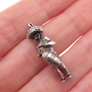 May include: A silver charm depicting a figure wearing a wide-brimmed hat with the word NASSAU embossed on the brim. The figure is shown playing a drum held in front of the body, with a small loop at the top for attachment to a bracelet or necklace.