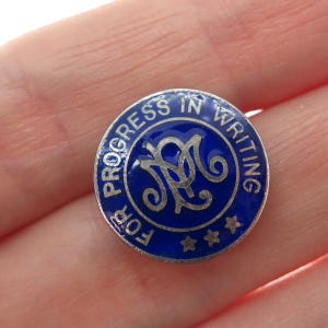 May include: A round, blue and silver pin with the words "PROGRESS IN WRITING" around the edge. The center features a stylized monogram. The pin has three small stars at the bottom. The pin is a vintage item.