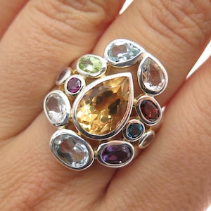 May include: A silver ring featuring a large, teardrop-shaped, golden-yellow gemstone surrounded by various smaller gemstones in shades of blue, green, purple, and red. The ring has a cluster design with a polished finish.