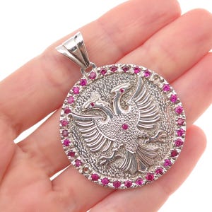 May include: A circular silver pendant featuring a relief of a double-headed eagle in the center. The border of the pendant is set with numerous small, round, vibrant pink gemstones. The metal surface has a textured, matte finish throughout.