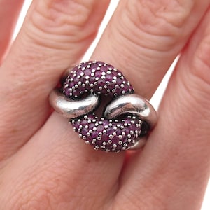 May include: A silver ring featuring two interlocking links, with the top and bottom sections densely encrusted with small, round, deep pinkish-red gemstones. The metal has a polished, slightly darkened finish, creating a bold, textured appearance.
