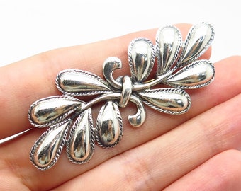 May include: Silver tone brooch with a stylized leaf design. The leaves are oval shaped and have a textured surface.