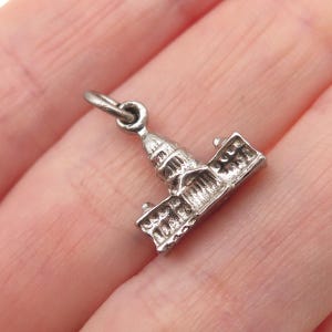 May include: A small, silver-toned charm in the shape of a building with a domed roof. The charm has intricate architectural details and a loop for attaching to a bracelet or necklace. The charm is held in a person's hand.