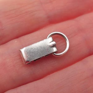 May include: A small, rectangular silver-toned metal charm featuring a smooth, flat surface and a rounded jump ring attached to one end. The piece has a polished finish and a simple, functional design, showing a clean, metallic appearance throughout.