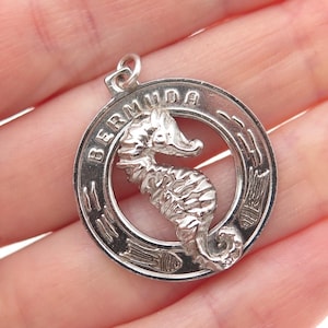 May include: A silver-toned pendant featuring a seahorse within a circular frame. The word "BERMUDA" is inscribed around the outer edge of the frame. The pendant is likely made of sterling silver and is designed to be worn as jewelry.