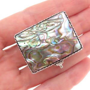 May include: A rectangular brooch featuring a polished abalone shell inlay with iridescent swirls of green, purple, and blue. The shell is set within a silver-toned metal frame that has a twisted rope-style border and a pin mechanism on the back.