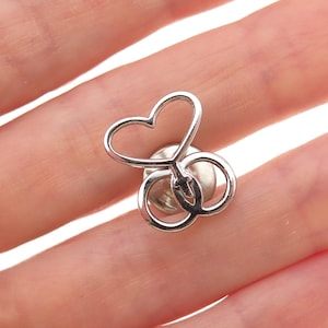 May include: A silver-toned pin with a heart and two interlocking circles design. The pin is a small, decorative accessory, likely for clothing or accessories. The heart shape is open, and the circles are intertwined, creating a unique and elegant look.