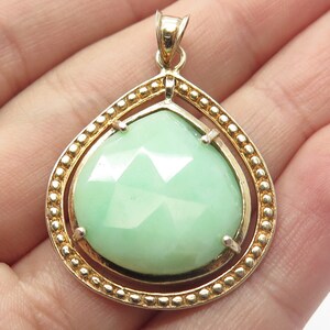 May include: A teardrop-shaped pendant with a green gemstone set in a gold-toned frame. The frame has a beaded edge.