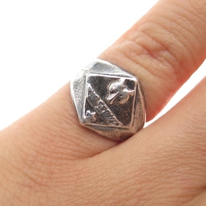 May include: A silver-toned ring with a square face featuring a detailed emblem. The emblem includes a bird, a feather, and other symbols. The ring is worn on a finger, showcasing its design and craftsmanship.