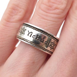 May include: A silver ring with the word "Yichud" engraved on it. The ring has a decorative border and Star of David symbols. The ring is worn on a finger.