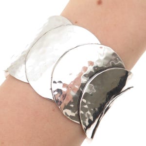May include: A silver-toned bracelet with a hammered texture. The bracelet is made of multiple, overlapping, crescent-shaped metal pieces. The bracelet is worn on a wrist.