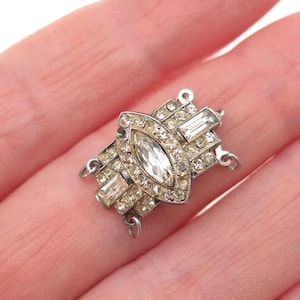 May include: Silver-toned vintage jewelry clasp with a central marquise-shaped clear crystal surrounded by smaller clear crystals. Rectangular and round clear crystals are set around the central design. The clasp has small loops on each end.
