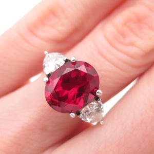 May include: A silver ring featuring a large, oval-cut, deep red gemstone as the centerpiece, flanked by two smaller, pear-shaped, clear gemstones. The ring's setting is silver, with small prongs securing the stones.
