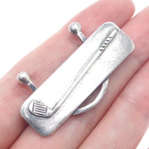 May include: Sterling silver golf club tie tack. The tie tack features a silver golf club with a ball on each end.