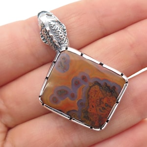 May include: A rectangular pendant featuring a polished, multi-colored agate stone with swirling patterns of orange, brown, and purple, set in a silver frame. The top of the pendant is adorned with a detailed, textured silver fish-shaped bail.