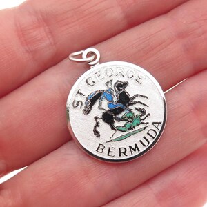 May include: A silver-toned round pendant with a detailed enamel design. The pendant features a black horse and rider, with blue and green accents, and the words "ST. GEORGE BERMUDA" around the edge. The pendant has a small loop at the top for a chain.