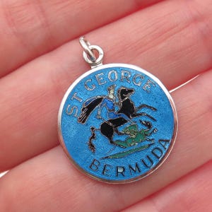 May include: A round, silver-toned pendant with a blue enamel background. The pendant features an image of a knight on a black horse, slaying a dragon. The words "ST. GEORGE" and "BERMUDA" are inscribed around the edge.