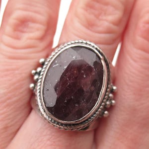 May include: A silver ring featuring a large, oval-cut, dark purple gemstone. The ring has a textured, braided border and small beaded accents on the sides, creating a vintage or antique aesthetic.