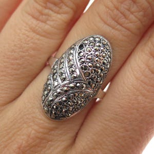 May include: Silver-toned ring with an oval face covered in small, dark, sparkling stones. The ring's design features a central pattern and is set against a light background.