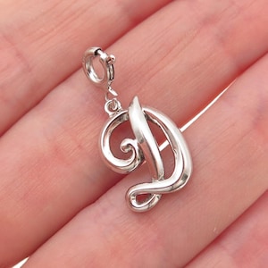 May include: A silver-colored charm in the shape of the letter "D". The charm has a decorative, cursive design with a loop at the top for attachment. The charm is shown against a light background.