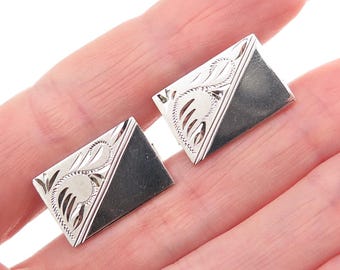 May include: A pair of silver cufflinks with a rectangular shape. Each cufflink features a diagonal design with engraved floral patterns and a dark gray section. The cufflinks are held in a hand.