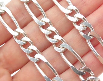 May include: Close-up of a silver chain with a Figaro link design. The chain is made of polished metal, with alternating elongated and oval links. The links are connected in a repeating pattern. The chain is displayed against a neutral background.