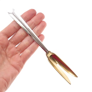 May include: A silver and gold-toned sugar tong with a decorative handle and a scoop-like design. The tongs are held in a hand against a white background. The handle has a detailed pattern.