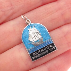May include: A silver-tone Bermuda charm with a blue enamel background. The charm features a white sailing ship and the text "BERMUDA DISCOVERED 1515". The charm has a small loop for attaching to a necklace or bracelet.