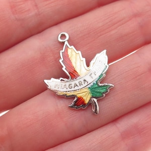 May include: A silver-toned maple leaf charm with enamel detailing. The leaf features red, yellow, and green sections, with the words "NIAGARA FALLS" on a white banner. The charm has a small loop at the top for attachment.
