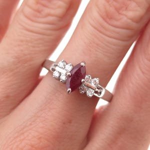 May include: A silver-toned ring featuring a central marquise-cut deep red gemstone flanked by two clusters of three small, clear, round-cut stones on each side, set on a polished metal band that encircles the finger.