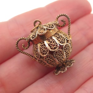 May include: A small, ornate gold-colored filigree urn with a hinged lid and two twisted rope handles. The urn has a floral base and a delicate, openwork design. The urn is likely a pendant or charm.