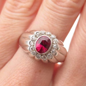 May include: A silver-toned ring featuring a central oval-cut deep red gemstone surrounded by a halo of twelve small, clear, round-cut stones. The band has a wide, polished metal design with vertical grooves extending from the central setting.