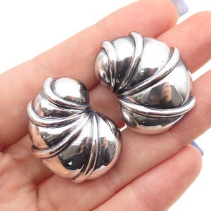 May include: A pair of polished silver earrings. Each earring has a rounded, shell-like design with curved, ridged details. The earrings are reflective, showing a mirrored surface.