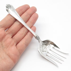 May include: A silver fork with an ornate design on the handle and tines. The fork is held in a hand, showing its size and detail.