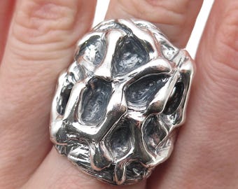 May include: A silver ring with an organic, textured design. The ring has a unique, irregular shape with a network of interconnected, rounded forms. The metal has a polished, reflective surface, and the ring is shown on a finger.