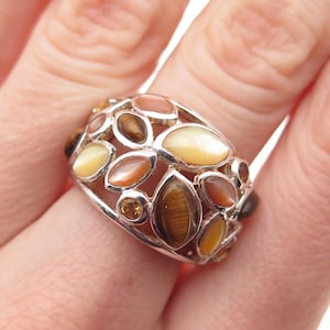 May include: A silver ring featuring a cluster of oval and teardrop-shaped gemstones in shades of brown, yellow, and orange. The ring has an openwork design, with the gemstones set within a silver band. The ring is a statement piece.