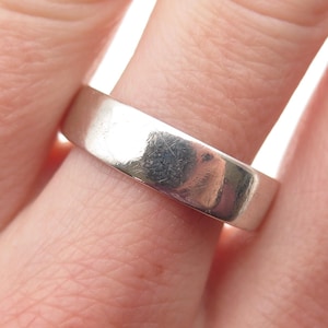 May include: A silver band ring with a flat, polished surface. The ring is worn on a finger and reflects light, showing minor wear and scratches. The ring is a simple, classic design.