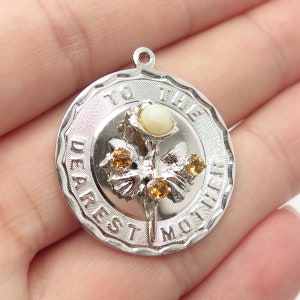 May include: Silver charm with a white and yellow flower design. The charm says "To the Dearest Mother".