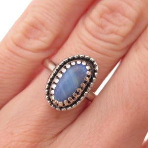May include: A silver ring featuring an oval-shaped, iridescent blue stone set in a scalloped, textured metal bezel. The band is smooth and metallic, holding the central stone securely in place with a decorative, notched edge surrounding the gem.
