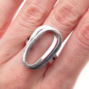 May include: A silver-toned ring featuring a large, open, elongated oval frame. The metal surface has a smooth, polished finish with subtle signs of wear, and the design consists of a continuous, hollow loop structure that sits prominently on the finger.