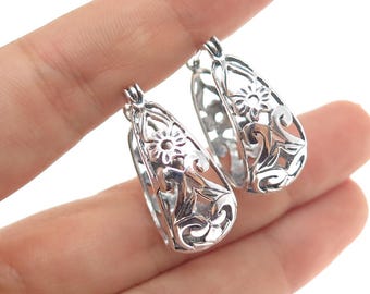 May include: A pair of silver-toned hoop earrings. Each earring features a floral and scrollwork design within a teardrop shape. The earrings are held up by a person's fingers.