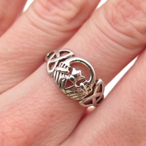 May include: A silver-toned Claddagh ring featuring a central heart held by two hands, topped with a crown, and flanked by intricate Celtic knotwork patterns on both sides of the band. The metal has a polished finish with visible engraved details.