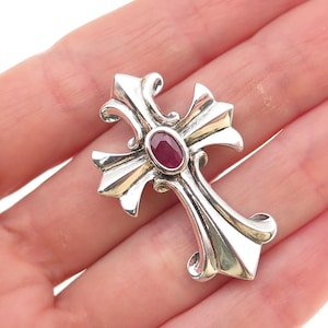 May include: A silver cross pendant with an oval, deep red gemstone at the center. The cross has ornate, curved arms and a polished finish, reflecting light. A classic religious symbol.