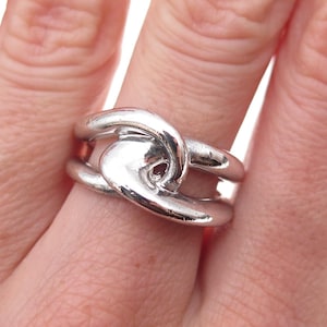 May include: A polished silver-tone ring featuring a continuous, fluid band that loops over itself in the center, creating an open, interconnected design with smooth, rounded edges and a reflective metallic finish that wraps around the finger.