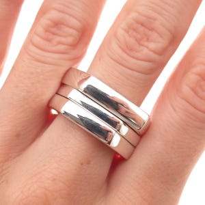 May include: A set of three stacked silver rings with a smooth, polished, and reflective surface. Each ring features a flat, wide band design with rounded edges, creating a uniform and cohesive appearance when worn together on a single finger.
