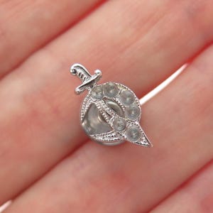 May include: A small, silver-toned brooch featuring a decorative design. The brooch has a central circular element with a series of smaller circles and a dagger-shaped accent. The brooch is likely made of metal and has intricate detailing.