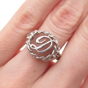 May include: A silver-toned ring featuring a central script letter D enclosed within a textured, rope-like circular frame. The metal has a polished finish on the letter and a rugged, uneven surface on the surrounding border of the ring band.