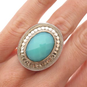 May include: An oval ring featuring a large, faceted turquoise-colored stone set in a gold-toned bezel. The stone is surrounded by a textured border of small silver-toned dots and a twisted rope pattern, all set within a polished silver-toned frame.